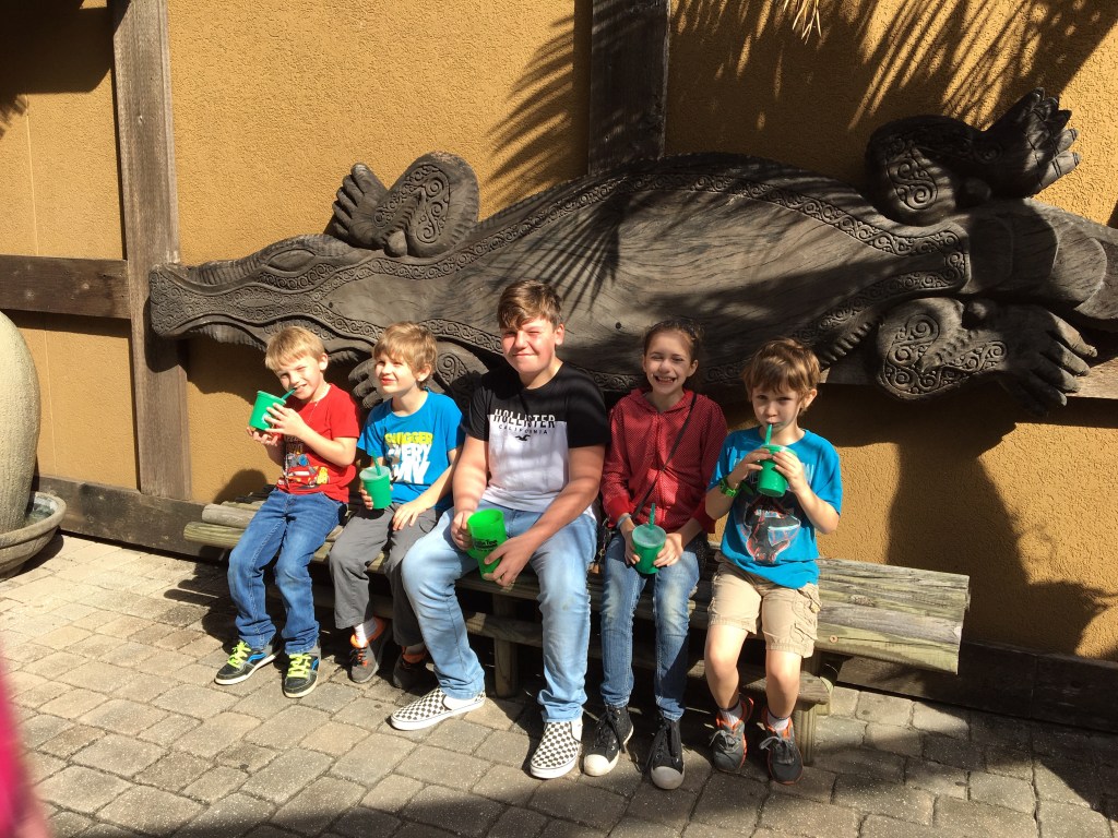 A group of five children sitting on a bench in front of a large carved wall decoration, each holding green cups.