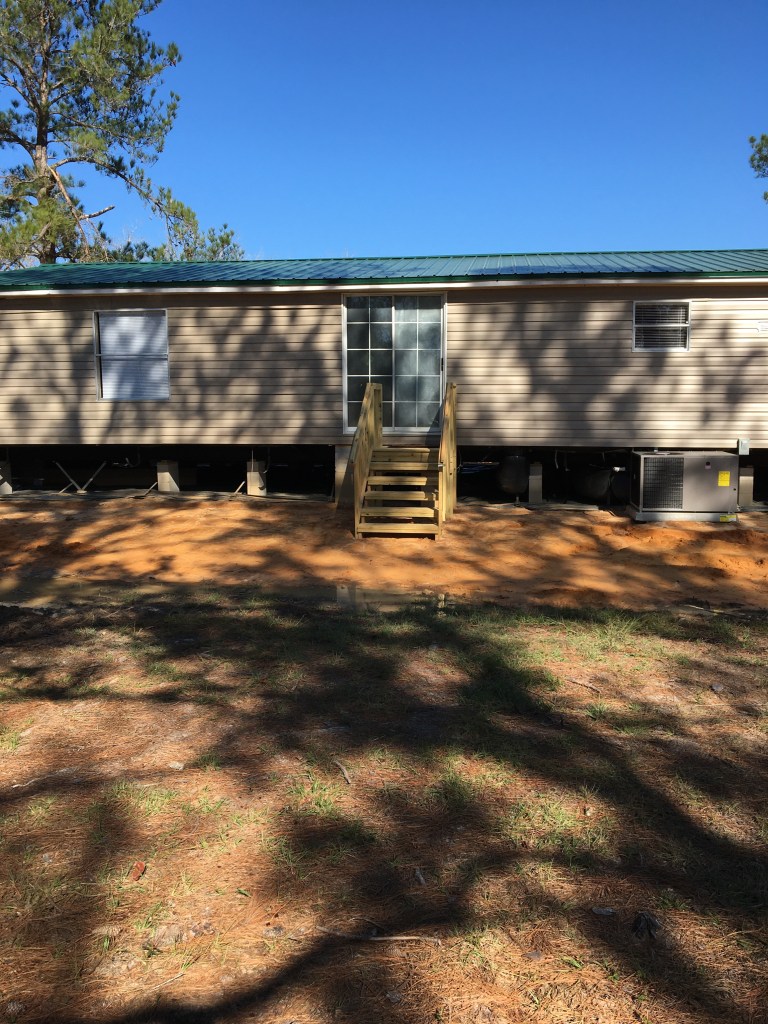 Front view of a mobile home with a wooden staircase, surrounded by pine trees and a clear blue sky.
